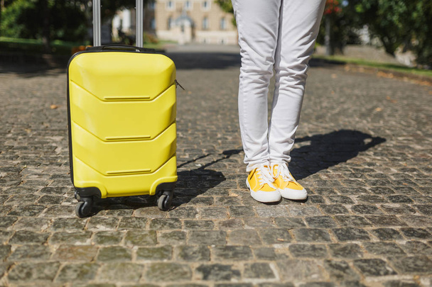 Περικοπεί εξωτερική εικόνα του ταξιδιώτη τουριστικά γυναίκα πόδια σε κίτρινο καλοκαίρι casual ρούχα με βαλίτσα στο δρόμο στην πόλη. Κορίτσι ταξίδια στο εξωτερικό για να ταξιδέψετε σε φυγή τα Σαββατοκύριακα. Τουρισμού ταξίδι ζωής - Φωτογραφία, εικόνα