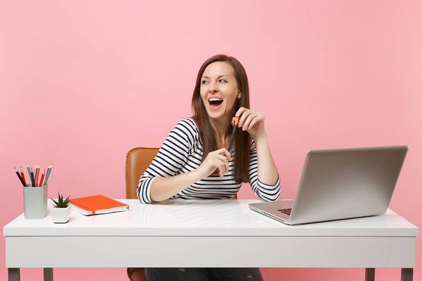Młoda Wesoła kobieta z otwarte usta trzymać ołówek właśnie siedzieć na bok pracy biały biurko z nowoczesnym laptopem na białym tle na tle pastelowy róż. Osiągnięcia firmy kariera koncepcja. Miejsce - Zdjęcie, obraz