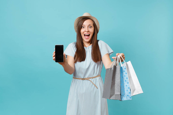 Portrait of woman in summer dress, straw hat holding packages bags with purchases after shopping, mobile cellphone with empty screen isolated on blue pastel background. Copy space for advertisement - Фото, изображение