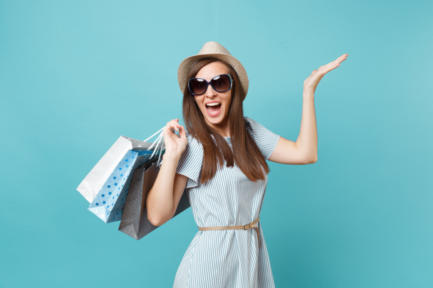 Portrait fashionable attractive happy woman in summer dress, straw hat, sunglasses holding packages bags with purchases after shopping isolated on blue pastel background. Copy space for advertisement - 写真・画像