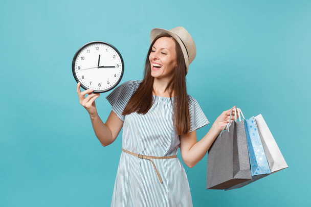 Portrait beautiful young caucasian woman in summer dress, straw hat holding packages bags with purchases after shopping, round clock isolated on blue pastel background. Copy space for advertisement - Fotografie, Obrázek