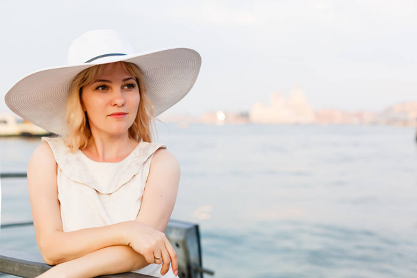 Портрет моди. Усміхнена блондинка в солом'яному капелюсі сидить на морському фоні
 - Фото, зображення