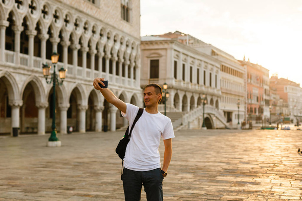 Joven hombre guapo mirando el teléfono en la Plaza San Marco en Venecia al amanecer, Italia, vistiendo camiseta blanca
 - Foto, Imagen