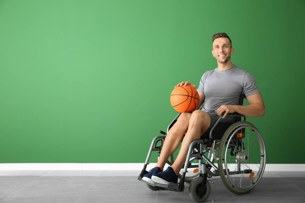 Νεαρός μπασκετμπολίστας που κάθεται σε αναπηρικό αμαξίδιο σε χρώμα τοίχο - Φωτογραφία, εικόνα