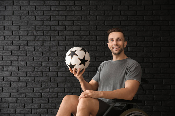 Young man with ball sitting in wheelchair against dark brick wall - Foto, imagen