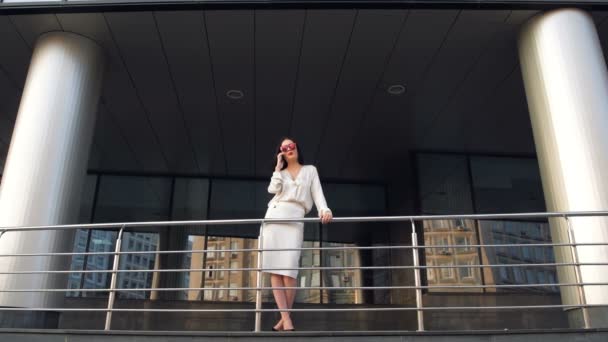 Beautiful elegant brunette businesswoman with mobile phone talking, smiling and laughing in front of skyscraper building where she works - Footage, Video