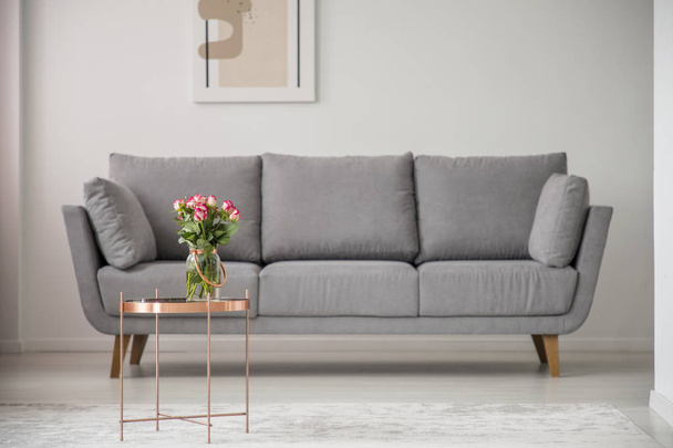 Flowers on copper table in front of grey sofa in bright living room interior with poster. Real photo - Valokuva, kuva