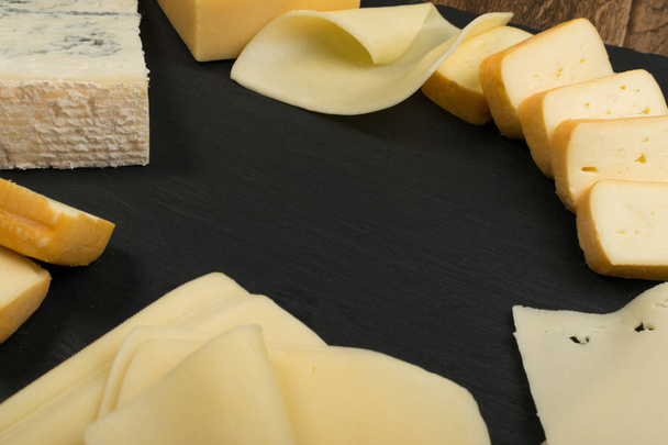 Sýrový nářez s plátky Limburger, Herve sýrem nebo Reblochon a modré Gorgonzola, Roquefort nebo Stilton. Sýr kurz s kombinací žluté a modré sýry pohled shora na černém kamenná deska pozadí - Fotografie, Obrázek