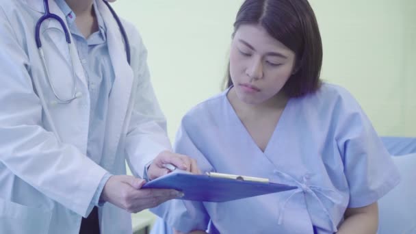 Magnifique médecin asiatique intelligent et patient discutant et expliquant quelque chose avec presse-papiers dans les mains du médecin tout en restant sur le lit du patient à l'hôpital. Médecine et soins de santé concept
. - Séquence, vidéo