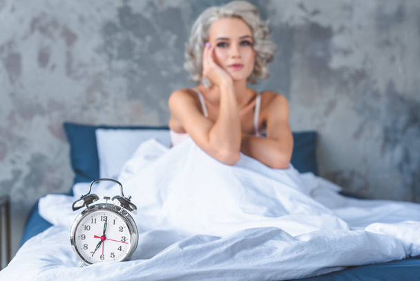 ült az ágyban vonzó fiatal nő borított fehér takaró vintage alarm Clock feküdt előtérben - Fotó, kép