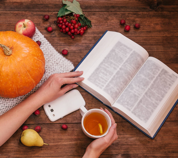 Vue de dessus de la main de la fille sur une table, une citrouille, des pommes et un livre, téléphone sur une table en bois
. - Photo, image