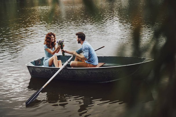 handsome young man giving bouquet to beautiful girlfriend while enjoying romantic date on lake in boat  - Photo, Image