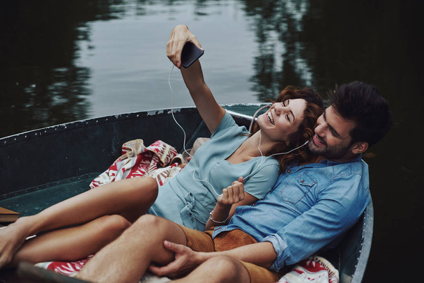 beautiful young couple listening music and taking selfie while sitting in boat outdoors - Foto, Bild