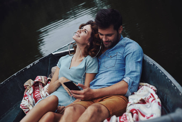 beautiful young couple listening music while sitting in boat outdoors - Zdjęcie, obraz