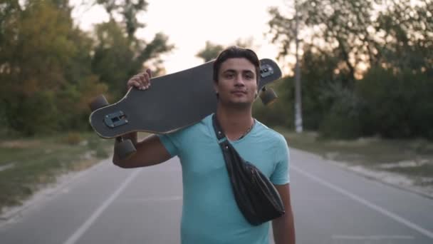 Jong gemengd ras hipster man met skateboard wandelen in stadspark bij zonsondergang - Video