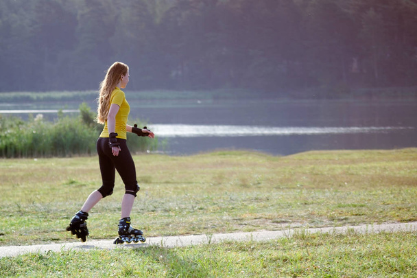 Mujer pelirroja joven, vestida con ropa deportiva amarilla, patinando cerca del lago a primera hora de la mañana
. - Foto, Imagen