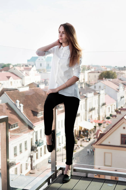 Joven mujer atractiva y elegante en camisa blanca y pantalones negros en el lugar al aire libre o plataforma de observación. Ciudad vieja en el fondo
. - Foto, imagen