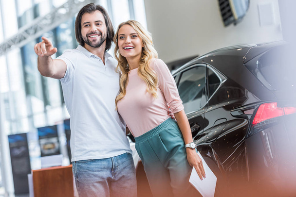 smiling couple with catalog buying car at dealership salon - Foto, afbeelding