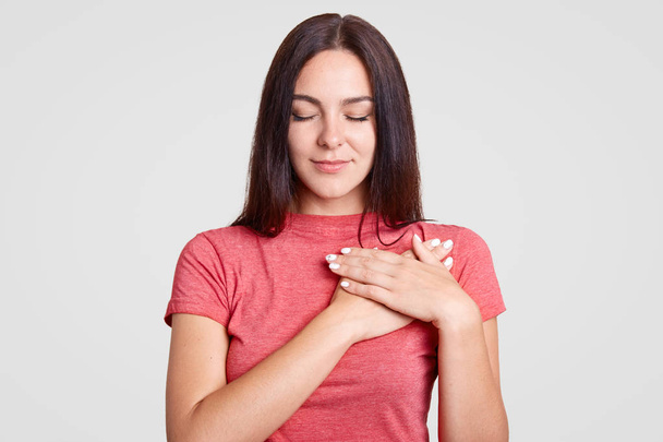 Calm brunette female with closed eyes, keeps both palms on heart, feels gratitude, being touched by something, dressed in casual pink t shirt, isolated over white background. Body language concept - Photo, Image