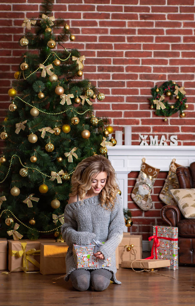 beautiful woman in knitted dress sits on her lap and holds a New Year's gift in her hands in the background of a luxurious decorated room with a Christmas tree - Zdjęcie, obraz