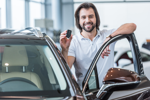 portrait of happy man with car key standing at new car in dealership salon - Фото, зображення