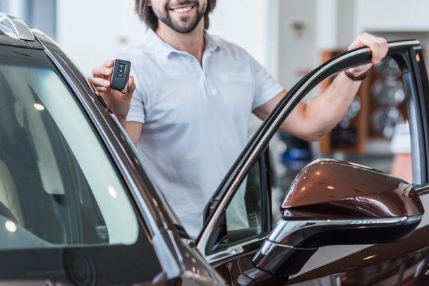 περικοπεί το πλάνο της χαμογελαστός άνθρωπος με αυτοκίνητο κλειδί στέκεται στο νέο αυτοκίνητο σε αντιπροσωπεία σαλόνι - Φωτογραφία, εικόνα