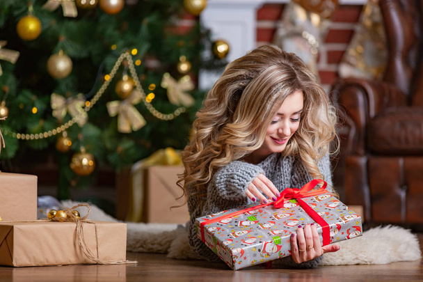 beautiful blonde lays on a warm rug and looks at a bright Christmas gift with red ribbon near the Christmas tree - Zdjęcie, obraz
