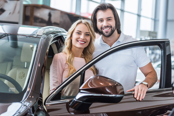 Portret van glimlachen paar staande op de nieuwe auto op de salon van de dealer - Foto, afbeelding