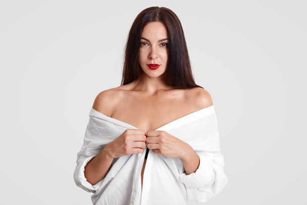 Horizontal shot of sexual female model with dark hair, unbuttons shirt, being half naked, poses for making shots to fulfill her portfolio, looks mysteriously at camera, has make up and red lips - 写真・画像