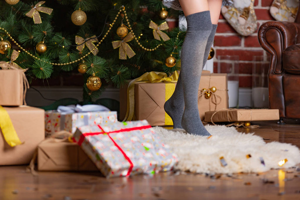woman stands on tiptoe in a warm gaiters on a fur rug in the room near the Christmas tree with presents - Фото, зображення