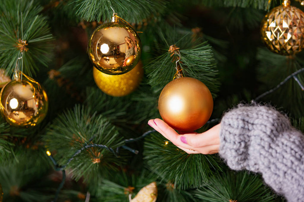 female hand touches the golden ball that hangs on the Christmas tree in the room. Close-up - Foto, Bild