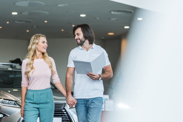 young couple with catalog buying car at dealership salon - Foto, Imagen