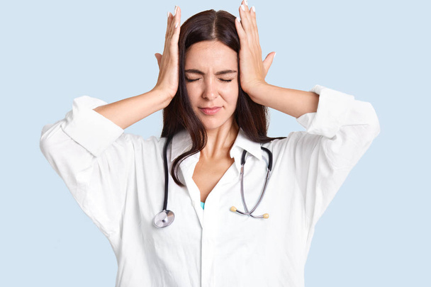 Horizontal shot of overworked female therapist has headache after hard working day and examining many patients, has phonendoscope on neck, isolated over light blue background, suffers from migraine - Foto, imagen