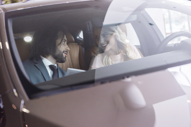 dealer salon verkoper en lachende vrouwelijke klant zitten in auto in auto salon - Foto, afbeelding