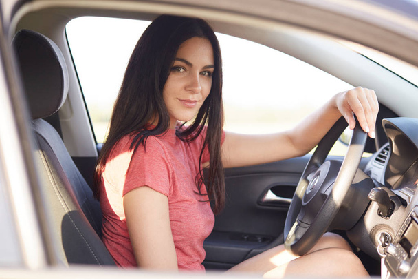 Young gorgeous brunette lady drives car alone, looks confidently at camera, keeps hand on wheel, likes travelling by automobile, enjoys high speed, has appealing look. Woman covers distance. - Фото, изображение