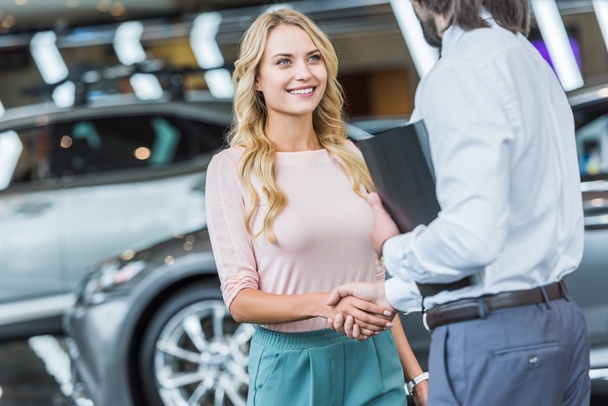 partial view of dealership salon shop assistant and smiling female customer shaking hands in auto salon - Fotó, kép