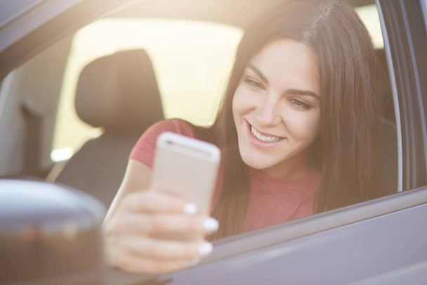 Glad European female with long dark hair, happy to recieve message on smart phone, poses in car, stops on road, connected to wireless internet, has positive smile. Woman driver with cellular - Photo, image