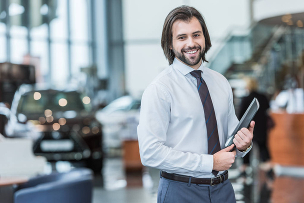 smiling seller with folder standing in dealership salon with cars on background - Zdjęcie, obraz