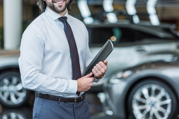 cropped shot of seller with folder standing in dealership salon with cars on background - Photo, Image