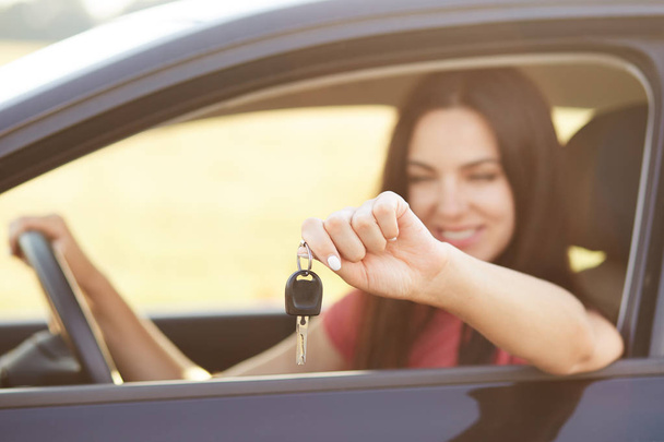Happy young brunette female holds key while sits in luxury automobile, glad to recieve expensive present from relatives, focus on keys, blurred background. People, transport, purchase concept - Фото, зображення