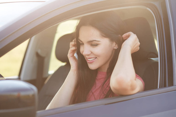 lächelnde brünette Fahrerin telefoniert mit einer engen Freundin, sitzt im Auto, hat zufriedene Mienen, diskutiert etwas, ist erfolgreiche Firmeninhaberin. Fahrerin mit modernem Gerät - Foto, Bild