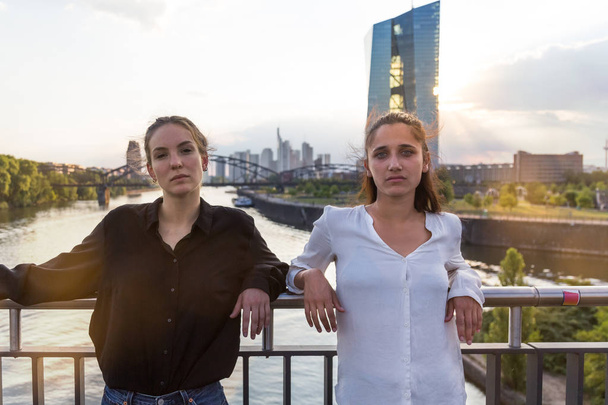 Deux jeunes femmes posant sur le pont de la ville sur la rivière avec la ligne d'horizon de la métropole en arrière-plan. Plan moyen
.  - Photo, image
