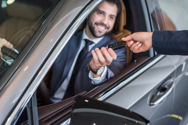 partial view of female seller giving car key to smiling businessman in new car at dealership salon - Fotó, kép