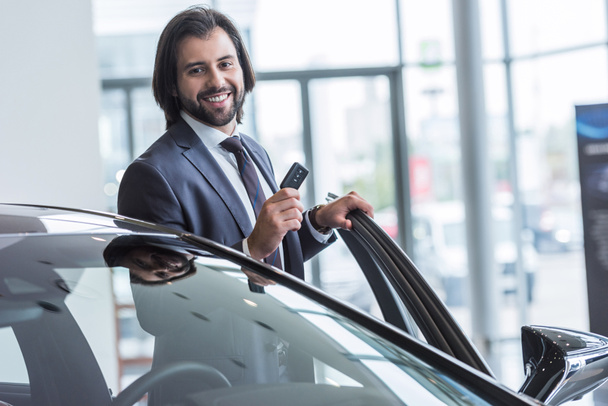 portrait of smiling businessman with car key standing at new car at dealership salon - Foto, imagen