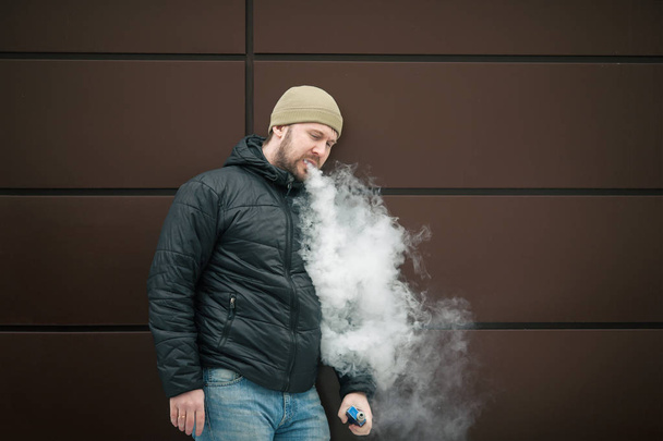 Vape man. Young bearded white guy in the cap and black jacket is smoking an electronic cigarette and letting off the steam opposite modern brown background. Lifestyle. - Foto, Imagem
