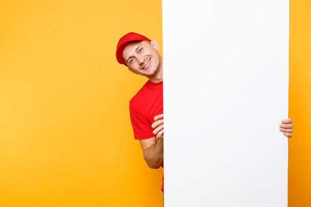Delivery man in red uniform isolated on yellow orange background. Male employee courier in cap, t-shirt holding big white empty blank billboard with copy space, place for text image. Service concept - Zdjęcie, obraz