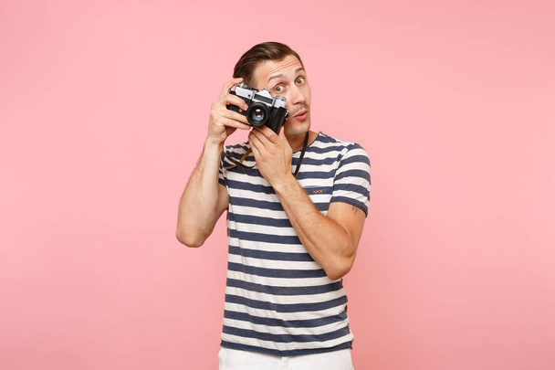 Портрет улыбающегося молодого фотографа в полосатой футболке фотографирует на ретро-винтажной фотокамере, изолированной на модном пастельно-розовом фоне. Люди искренние эмоции
 - Фото, изображение