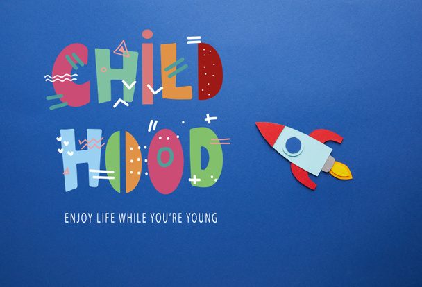 creative rocket on blue paper background with "childhood - enjoy life while you are young" lettering - Foto, Imagem