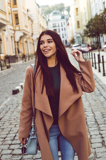 Podzim může být stylový. Krásná mladá žena v uhýbaje stylový kabát s úsměvem stoje venku - Fotografie, Obrázek