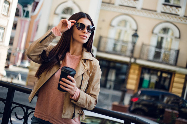 Всегда с нетерпением жду. Красивая молодая женщина в солнечных очках, держа чашку кофе и глядя прочь с улыбкой, стоя на улице
 - Фото, изображение